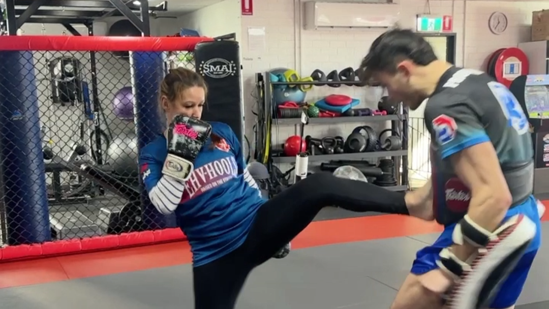 Person practicing martial arts in a gym with another person holding a kicking pad.