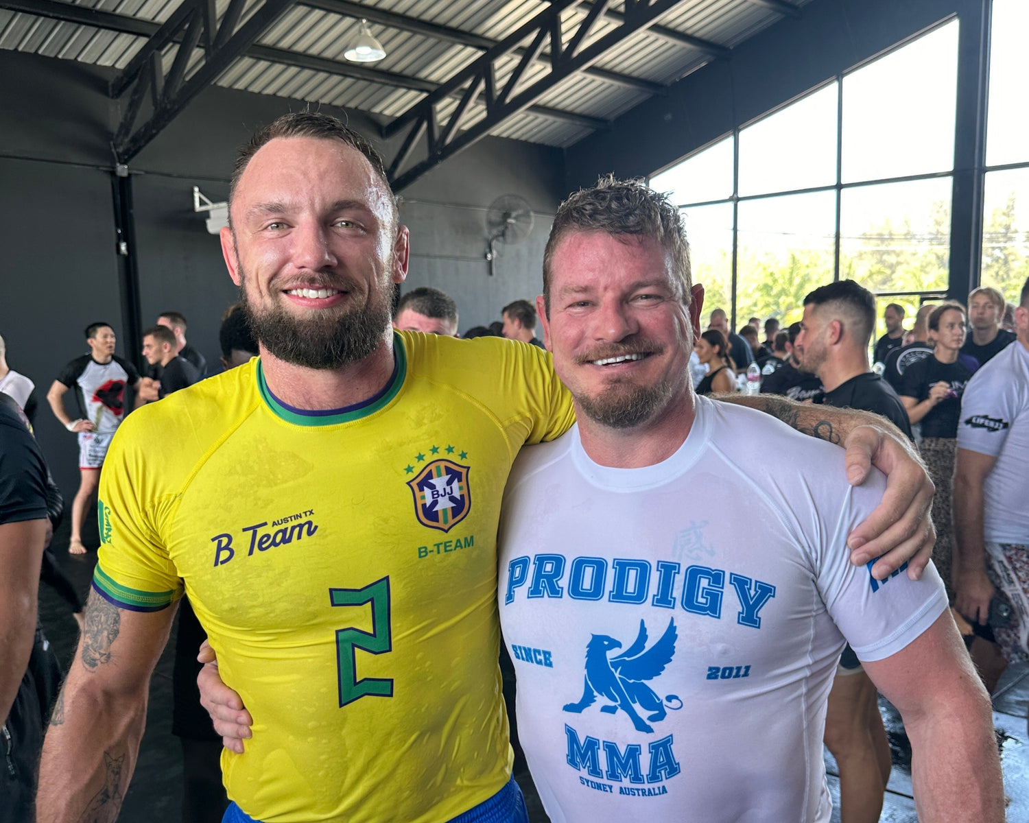 Two men standing together, one in a Brazil soccer jersey and the other in a 'Prodigy MMA' shirt, in an indoor setting with people in the background.