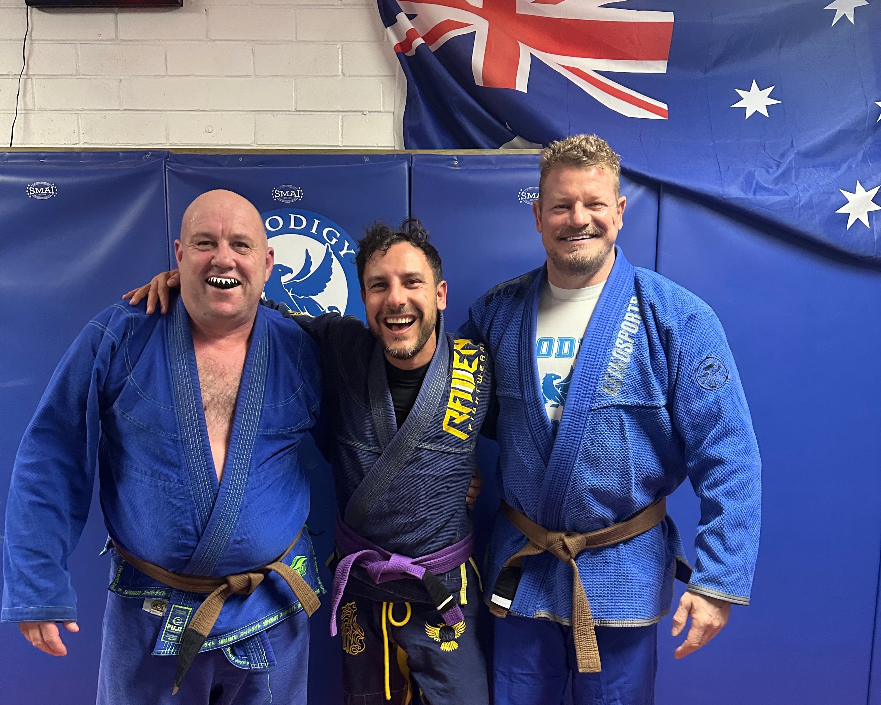 Three people in blue martial arts uniforms posing for a photo on a blue and orange mat with an Australian flag in the background.